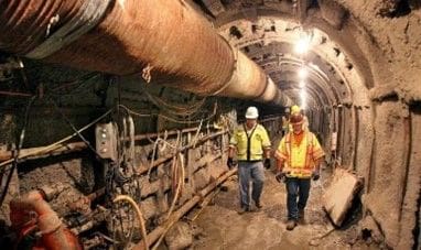 Two men in yellow jackets and hard hats walk through a tunnel.